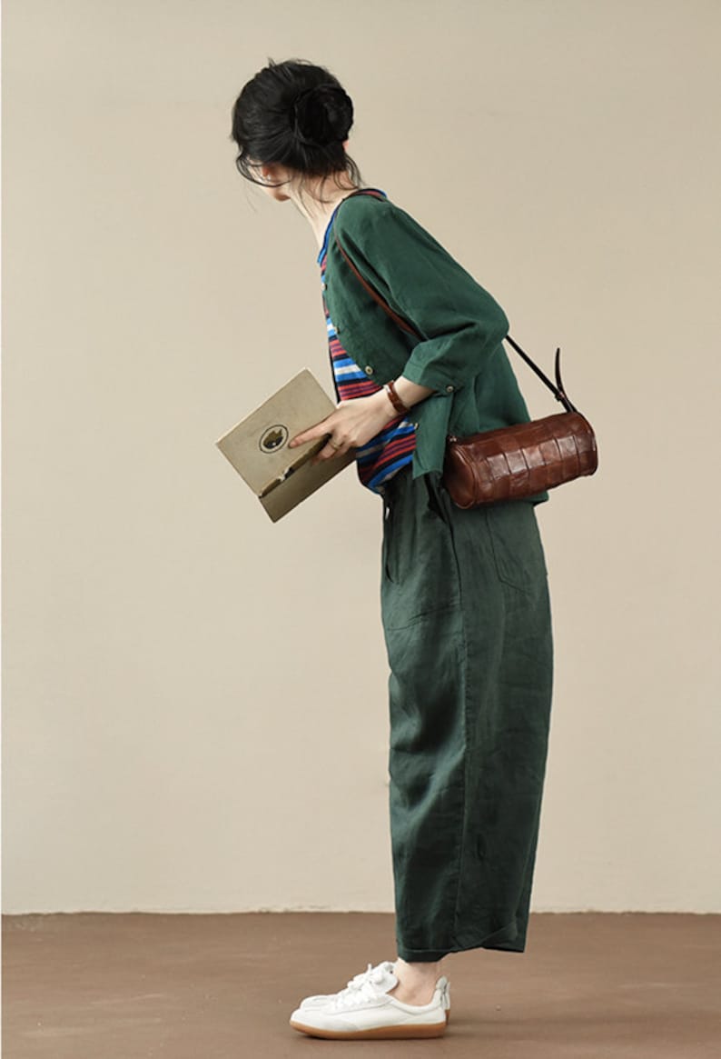 May include: A woman wearing a green linen shirt and pants with a brown leather crossbody bag. She is holding a book and wearing white sneakers.
