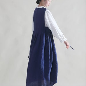Puede incluir: Un vestido de lino azul marino con una blusa blanca debajo. El vestido tiene una falda plisada y un lazo en la cintura. La modelo lleva un sombrero de paja con una cinta negra.