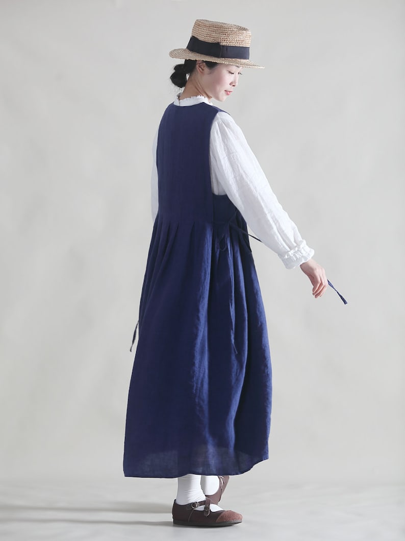 Puede incluir: Un vestido de lino azul marino con una blusa blanca debajo. El vestido tiene una falda plisada y un lazo en la cintura. La modelo lleva un sombrero de paja con una cinta negra.