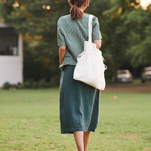 May include: A woman wearing a green and white gingham shirt and a long green skirt walks on a grassy field. She is carrying a white tote bag.