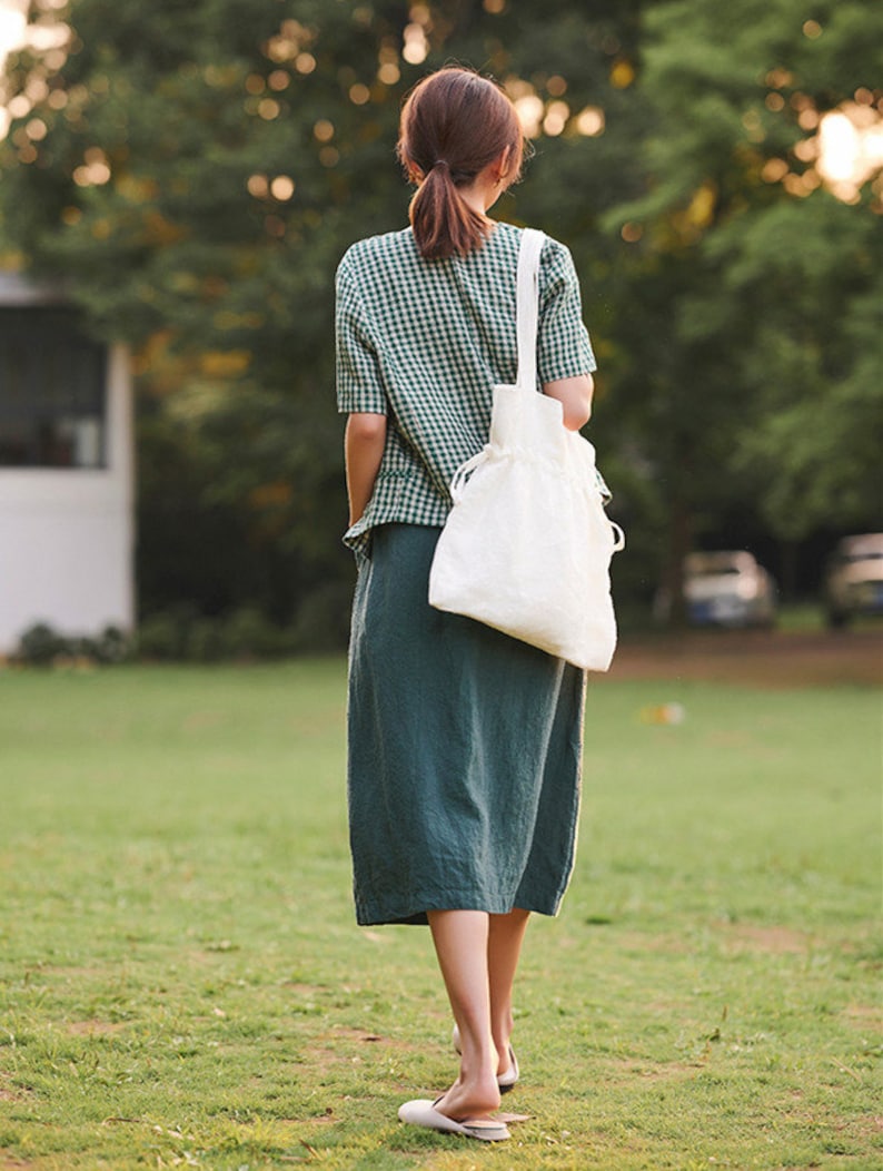May include: A woman wearing a green and white gingham shirt and a long green skirt walks on a grassy field. She is carrying a white tote bag.