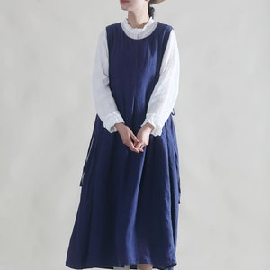 Puede incluir: Una mujer lleva un vestido de lino azul marino con una blusa blanca debajo. Lleva un sombrero de paja con una banda negra y zapatos marrones.