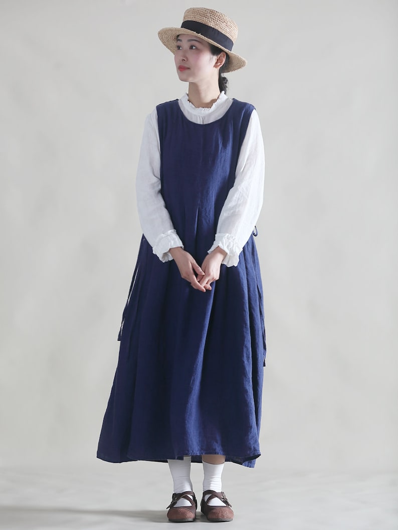 Puede incluir: Una mujer lleva un vestido de lino azul marino con una blusa blanca debajo. Lleva un sombrero de paja con una banda negra y zapatos marrones.