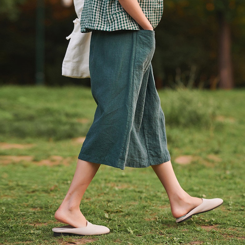 May include: A woman wearing a green linen wide-leg trouser with a white and green checked shirt. She is walking on a grassy lawn and wearing white slip-on shoes.