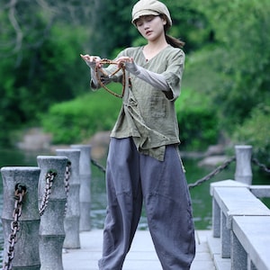 May include: A person wearing a sage green linen top, gray pants, and a matching cap, holding a beaded necklace. The outfit is loose-fitting and casual, with white sneakers. The background features a serene outdoor setting with water and greenery.