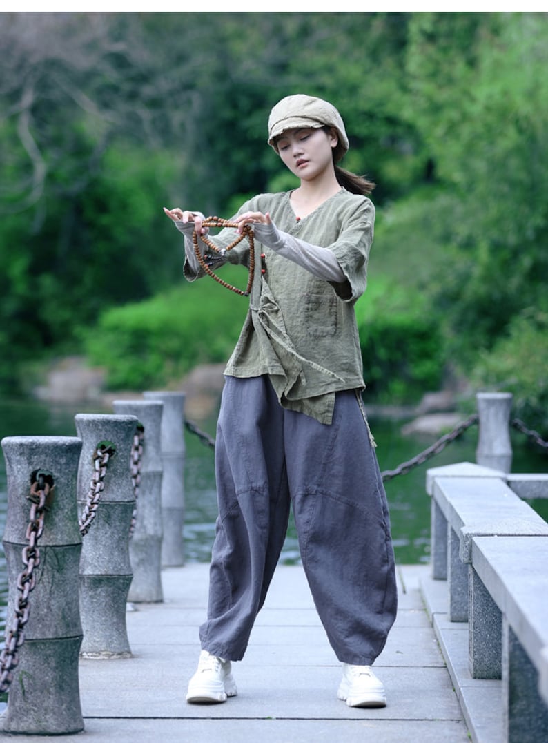 May include: A person wearing a sage green linen top, gray pants, and a matching cap, holding a beaded necklace. The outfit is loose-fitting and casual, with white sneakers. The background features a serene outdoor setting with water and greenery.