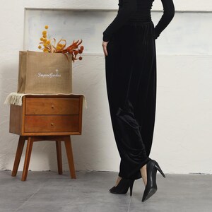 May include: A woman wearing black velvet pants and black heels. She is standing in front of a wooden nightstand with a burlap bag on top that says "Simpson Gardens".
