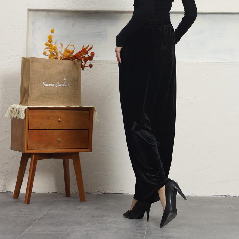 May include: A woman wearing black velvet pants and black heels. She is standing in front of a wooden nightstand with a burlap bag on top that says "Simpson Gardens".