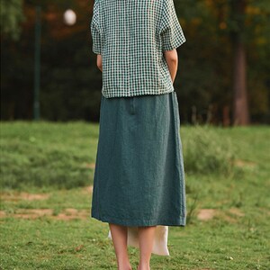 May include: A woman wearing a green and white gingham short-sleeved shirt and a long, dark green skirt.