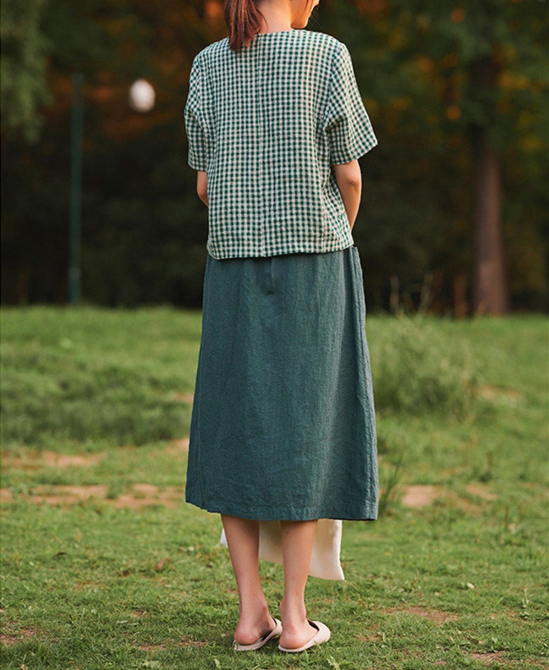 May include: A woman wearing a green and white gingham short-sleeved shirt and a long, dark green skirt.