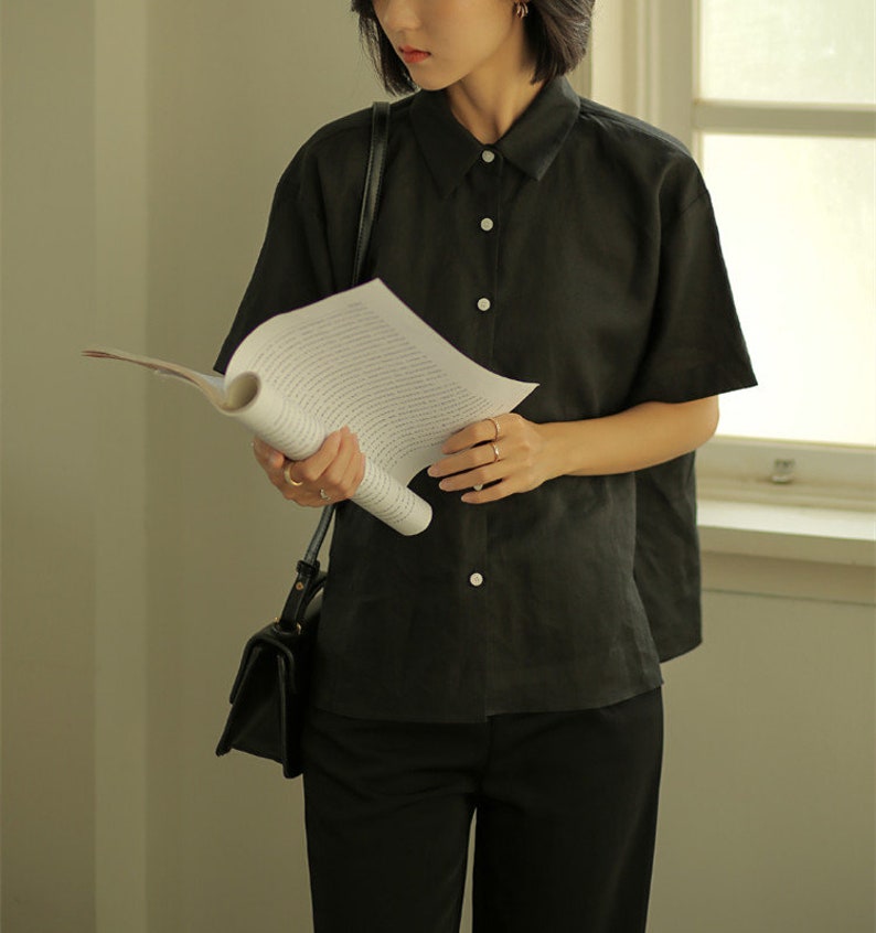 May include: A woman wearing a black short-sleeved collared shirt with buttons and black pants. She is holding a book and a black handbag.
