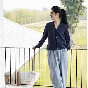 May include: A woman wearing a dark blue linen shirt with a button-up front and light blue linen pants. She is standing on a balcony overlooking a green field.
