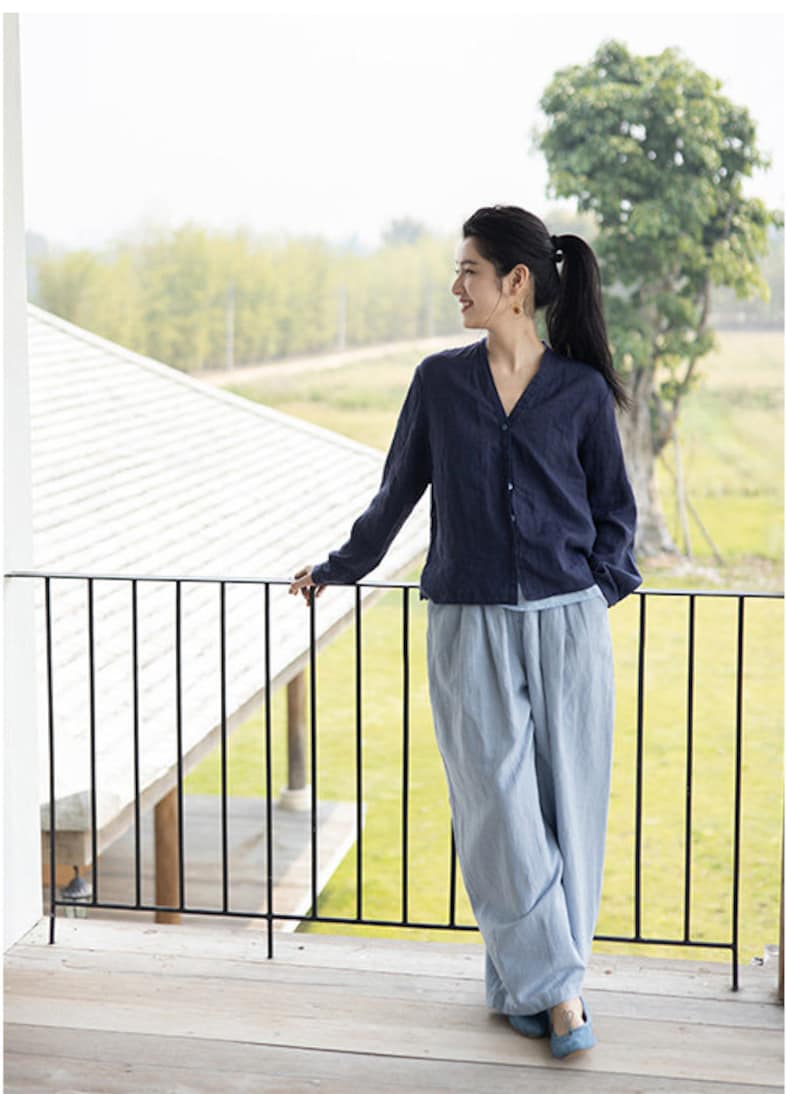 May include: A woman wearing a dark blue linen shirt with a button-up front and light blue linen pants. She is standing on a balcony overlooking a green field.