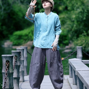 May include: A person wearing a light blue linen shirt with long sleeves and a mandarin collar, paired with dark gray, loose-fitting pants. The outfit is completed with a beige cap and white sneakers. A wooden beaded necklace is held up.