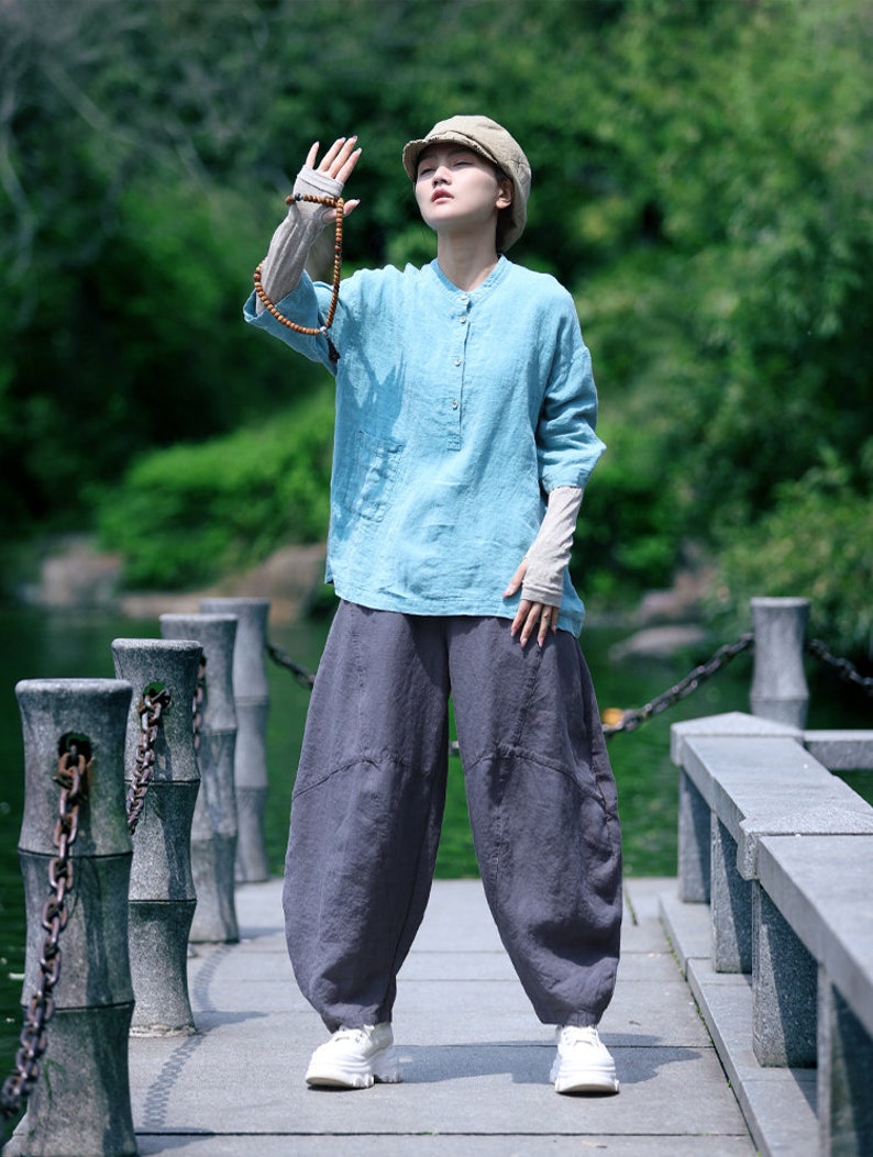 May include: A person wearing a light blue linen shirt with long sleeves and a mandarin collar, paired with dark gray, loose-fitting pants. The outfit is completed with a beige cap and white sneakers. A wooden beaded necklace is held up.