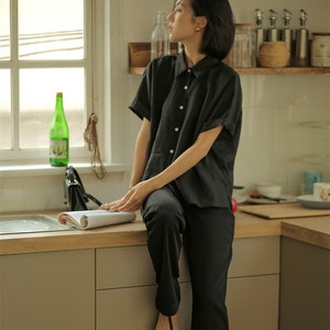 May include: A woman wearing a black short-sleeved collared shirt and black pants sits on a kitchen counter. She is holding a magazine and looking out a window.