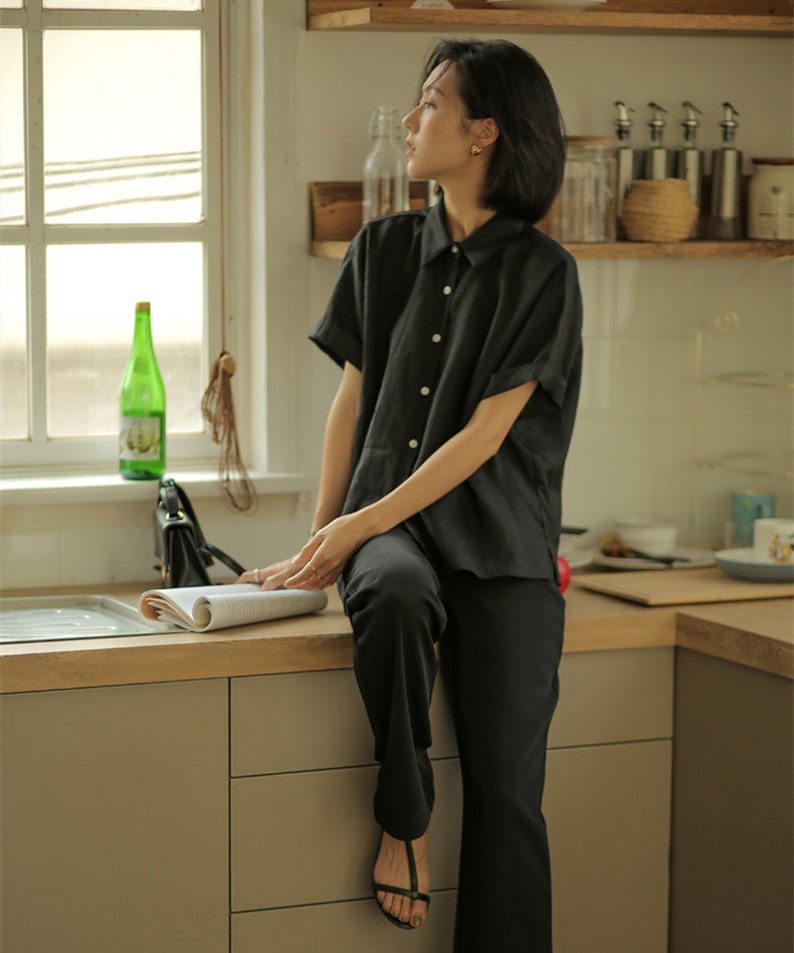 May include: A woman wearing a black short-sleeved collared shirt and black pants sits on a kitchen counter. She is holding a magazine and looking out a window.