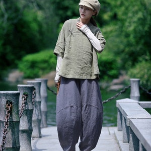 May include: An olive green linen top with short sleeves and a light gray long-sleeved shirt underneath. The outfit includes gray harem pants and white platform sneakers. A beige cap completes the look, suitable for casual wear.