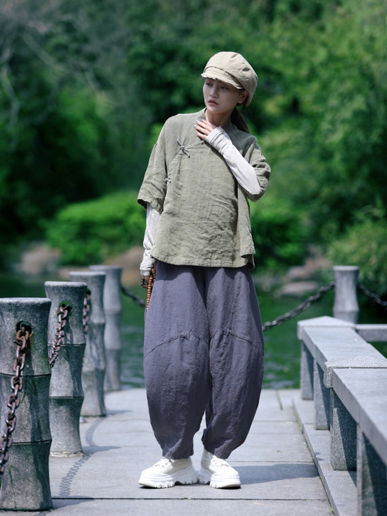 May include: An olive green linen top with short sleeves and a light gray long-sleeved shirt underneath. The outfit includes gray harem pants and white platform sneakers. A beige cap completes the look, suitable for casual wear.