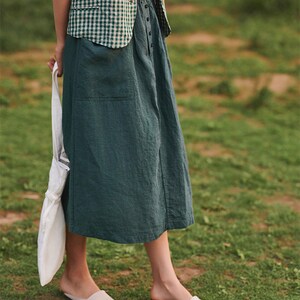 May include: A woman wearing a green and white checked shirt and a dark green skirt with a white tote bag. She is wearing white slip-on shoes and standing on a grassy surface.