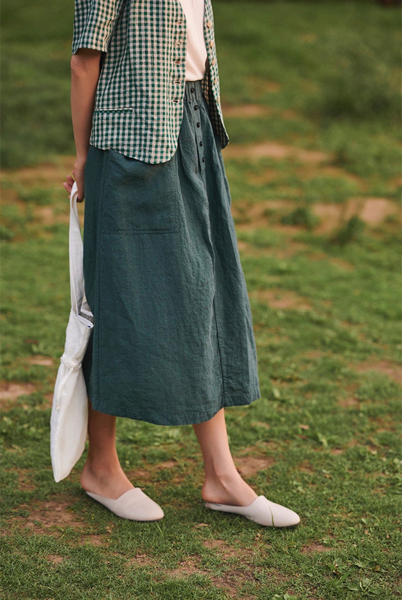 May include: A woman wearing a green and white checked shirt and a dark green skirt with a white tote bag. She is wearing white slip-on shoes and standing on a grassy surface.