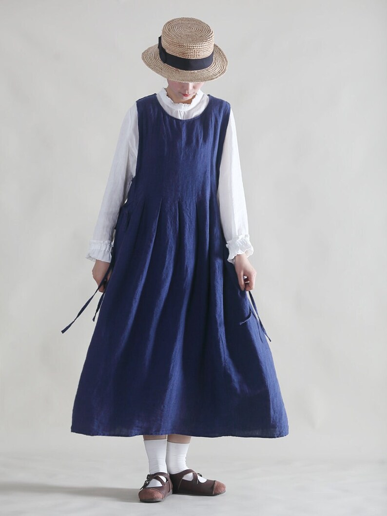 Puede incluir: Un vestido de lino azul marino sin mangas con una falda plisada. El vestido tiene tirantes ajustables que se atan a los lados. La modelo lleva una blusa blanca debajo del vestido y un sombrero de paja con una cinta negra.