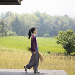 May include: A woman wearing a purple and blue jacket and gray pants walks on a concrete path. She is holding a bundle of dried grass in her right hand. The path is in front of a field of green grass and trees.
