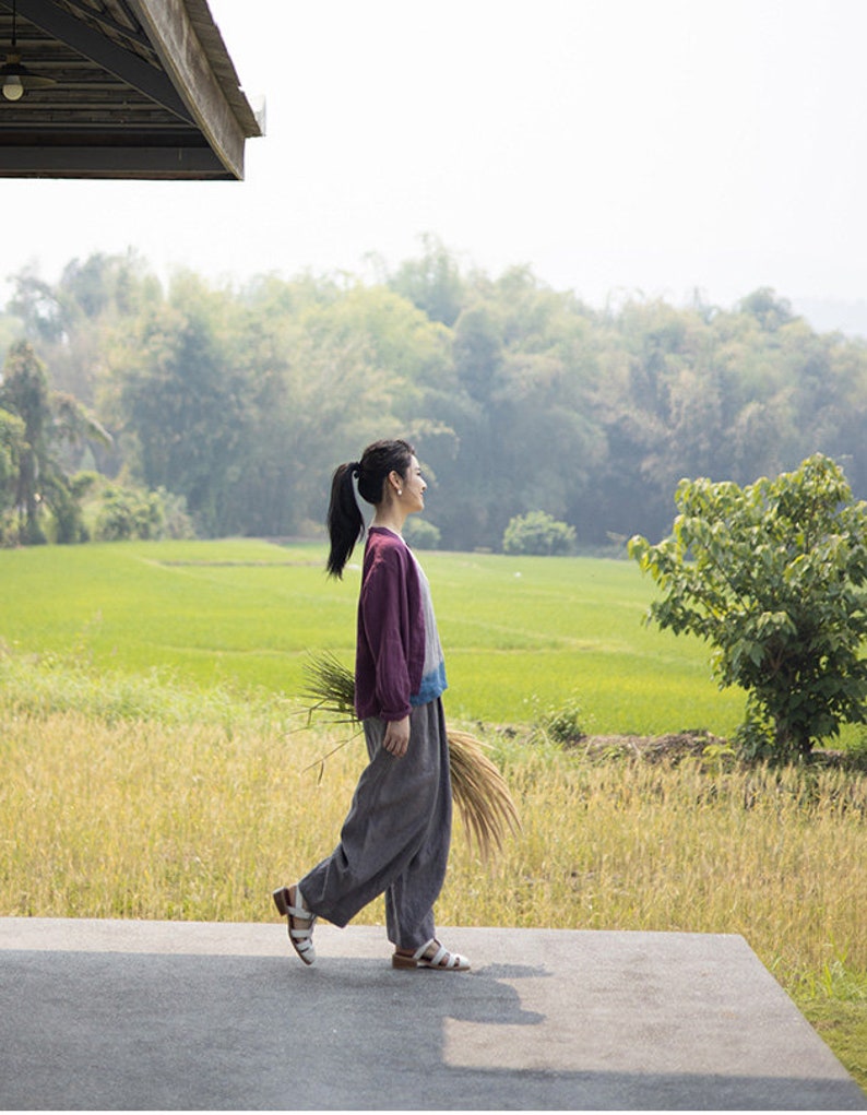 May include: A woman wearing a purple and blue jacket and gray pants walks on a concrete path. She is holding a bundle of dried grass in her right hand. The path is in front of a field of green grass and trees.