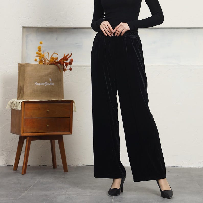 May include: Black velvet wide-leg pants with a high waist and a relaxed fit. The pants are shown on a model wearing a black turtleneck top and black heels.
