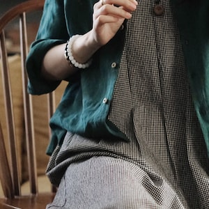 May include: A woman wearing a green linen shirt over a black and white gingham dress. She is wearing a white beaded bracelet and a necklace with a wooden pendant.