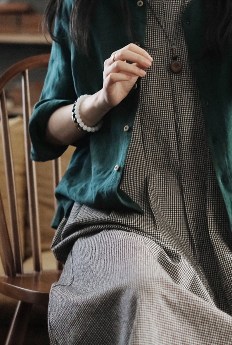 May include: A woman wearing a green linen shirt over a black and white gingham dress. She is wearing a white beaded bracelet and a necklace with a wooden pendant.