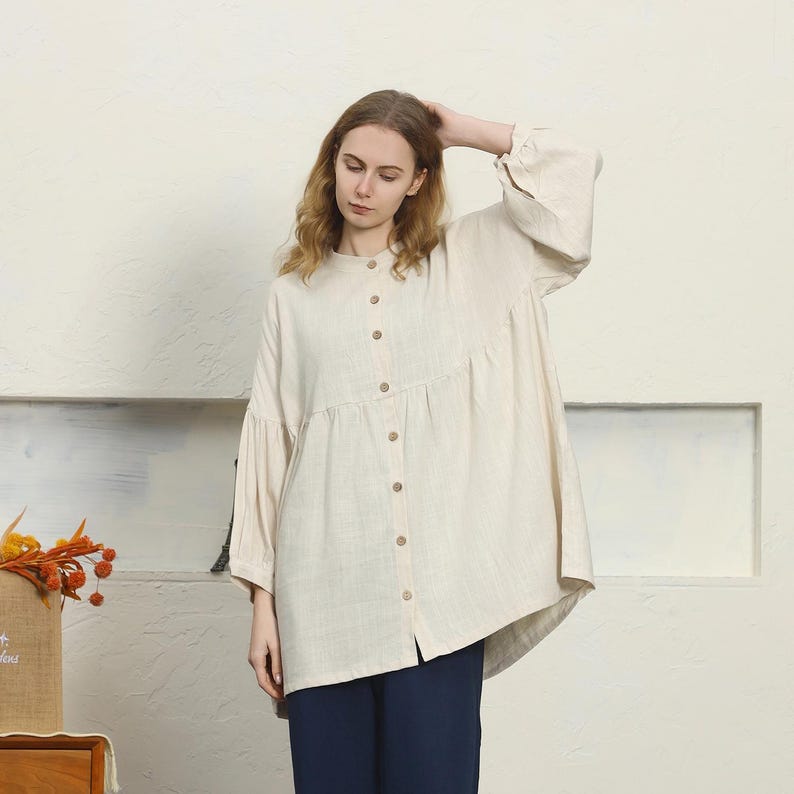 May include: A cream-colored button-down shirt with long sleeves and a rounded collar. The shirt features a gathered waist and wooden buttons. The model is wearing navy blue pants.