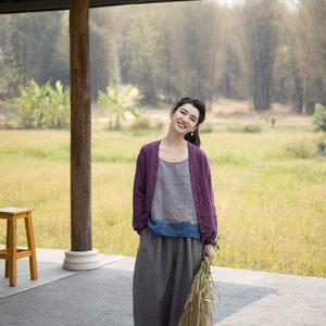 May include: A woman wearing a purple cardigan, a gray and blue top, and gray pants stands in front of a wooden structure. She is holding a bouquet of dried flowers and smiling at the camera.
