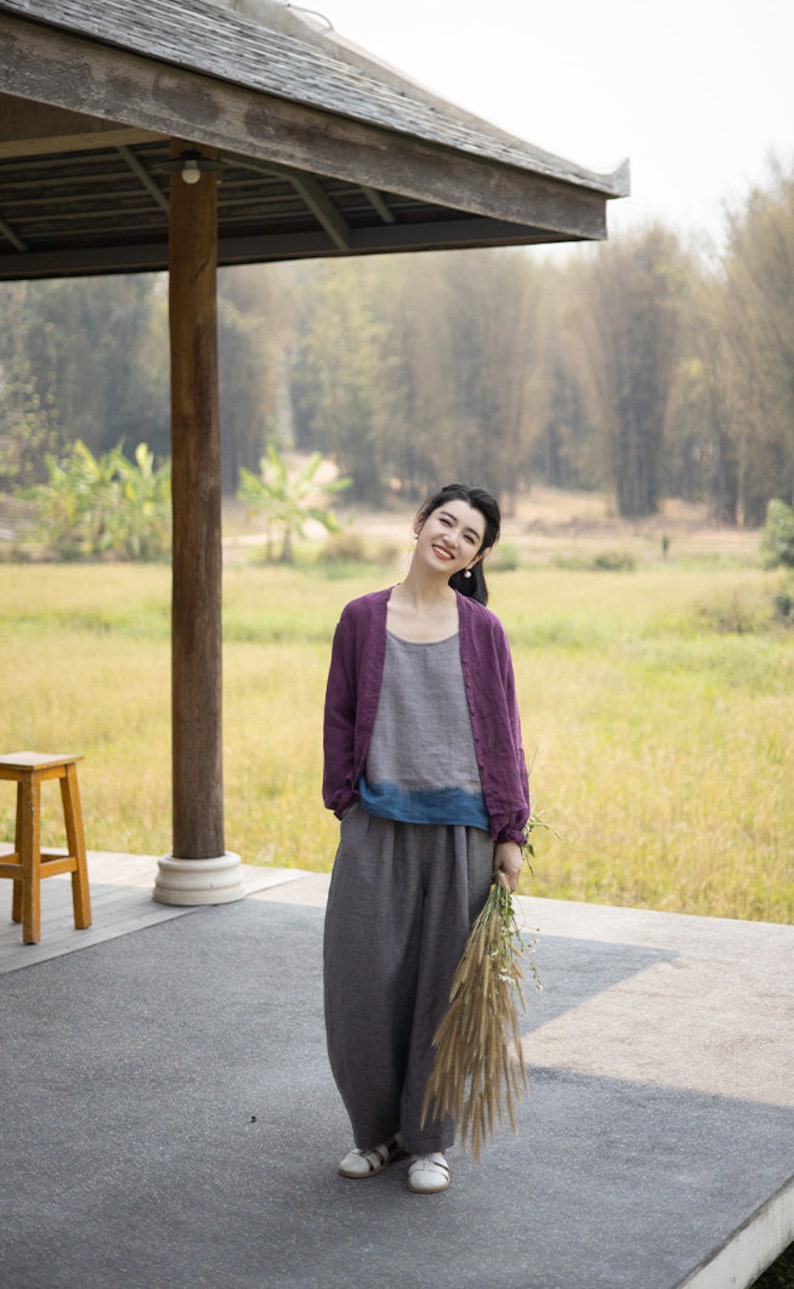 May include: A woman wearing a purple cardigan, a gray and blue top, and gray pants stands in front of a wooden structure. She is holding a bouquet of dried flowers and smiling at the camera.