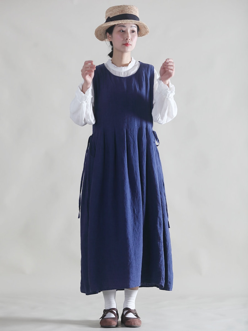 Puede incluir: Una mujer con un vestido de lino azul y una blusa blanca debajo. Lleva un sombrero de paja y zapatos marrones.