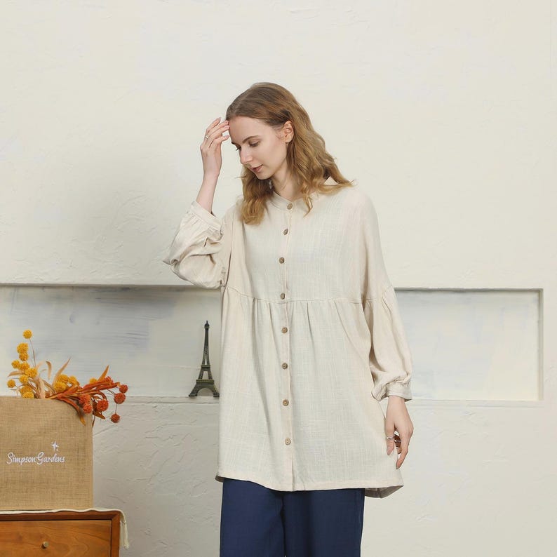 May include: A beige button-down tunic with long sleeves and a gathered waist. The tunic is paired with navy blue pants. A small Eiffel Tower statue and a box with the text "Simpson Gardens" are in the background.