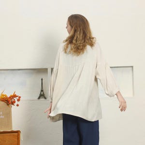 May include: A beige linen blouse with long, voluminous sleeves and a relaxed fit. The blouse is paired with navy blue pants. The model is facing away from the camera, with long, wavy blonde hair.