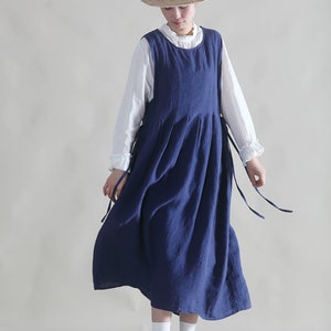 Puede incluir: Una mujer con un vestido de lino azul y una blusa blanca debajo. Lleva un sombrero de paja con una cinta negra y zapatos marrones.