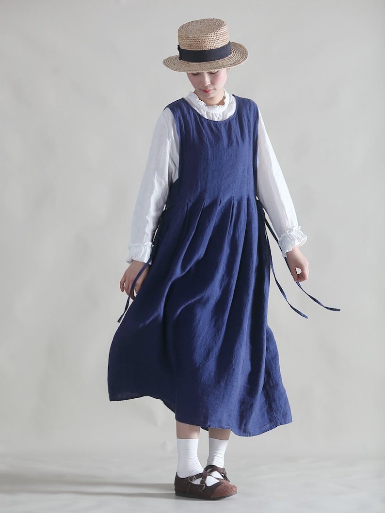 Puede incluir: Una mujer con un vestido de lino azul y una blusa blanca debajo. Lleva un sombrero de paja con una cinta negra y zapatos marrones.