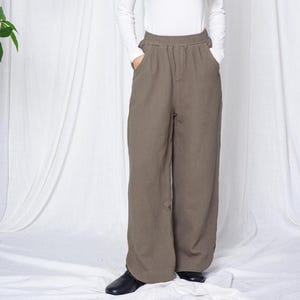 Könnte beinhalten: Eine Person trägt ein weißes Langarmshirt und eine olivgrüne Hose mit weitem Bein. Die Hose hat einen elastischen Bund und Taschen. Die Person trägt schwarze Schuhe. Der Hintergrund ist ein weißer Vorhang.