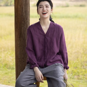 May include: A woman wearing a purple linen shirt and gray linen pants sits on a wooden stool. She is smiling and holding a bouquet of flowers.