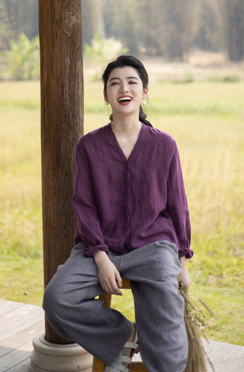 May include: A woman wearing a purple linen shirt and gray linen pants sits on a wooden stool. She is smiling and holding a bouquet of flowers.