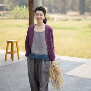 May include: A woman wearing a purple linen cardigan over a gray linen top and gray linen pants. She is holding a bouquet of dried flowers and smiling at the camera.