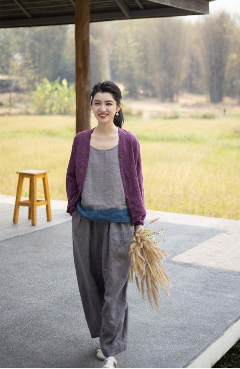 May include: A woman wearing a purple linen cardigan over a gray linen top and gray linen pants. She is holding a bouquet of dried flowers and smiling at the camera.