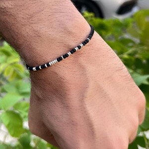 Könnte beinhalten: Schwarzes Perlenarmband mit silberfarbenen Akzenten. Das Armband besteht aus kleinen, runden schwarzen Perlen und silberfarbenen Perlen. Es wird am Handgelenk getragen, mit grünem Laub im Hintergrund.