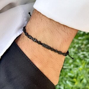 Può includere: Un braccialetto di perline nere con chiusura a nodo scorrevole. Il braccialetto è composto da piccole perline rettangolari infilate su un cordoncino nero. Il braccialetto è indossato su un polso, con una camicia bianca e pantaloni neri sullo sfondo.