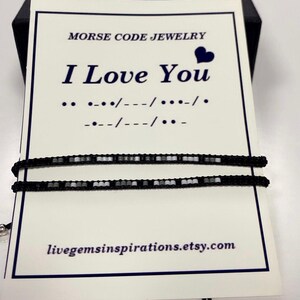 May include: A black beaded bracelet with white beads spelling out "I Love You" in Morse code. The bracelet is displayed on a white card with the message "Morse Code Jewellery" and the website address "livegemsinspirations.etsy.com".