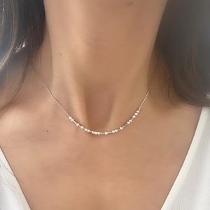 May include: A delicate silver necklace with small round beads. The necklace is worn by a person with a white shirt.