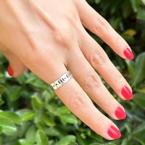May include: A silver ring with a unique design featuring a series of small, evenly spaced circles. The ring is worn on the middle finger of a hand with red nail polish.