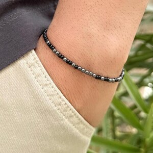 Könnte beinhalten: Ein Perlenarmband mit einer Mischung aus schwarzen und silbernen Perlen. Das Armband wird am Handgelenk getragen, mit einem grauen T-Shirt und einer beigen Hose im Hintergrund. Das Armband hat ein schlichtes, minimalistisches Design.